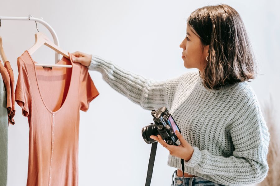 Mulher escolhendo roupas numa arara, com a câmera na mão